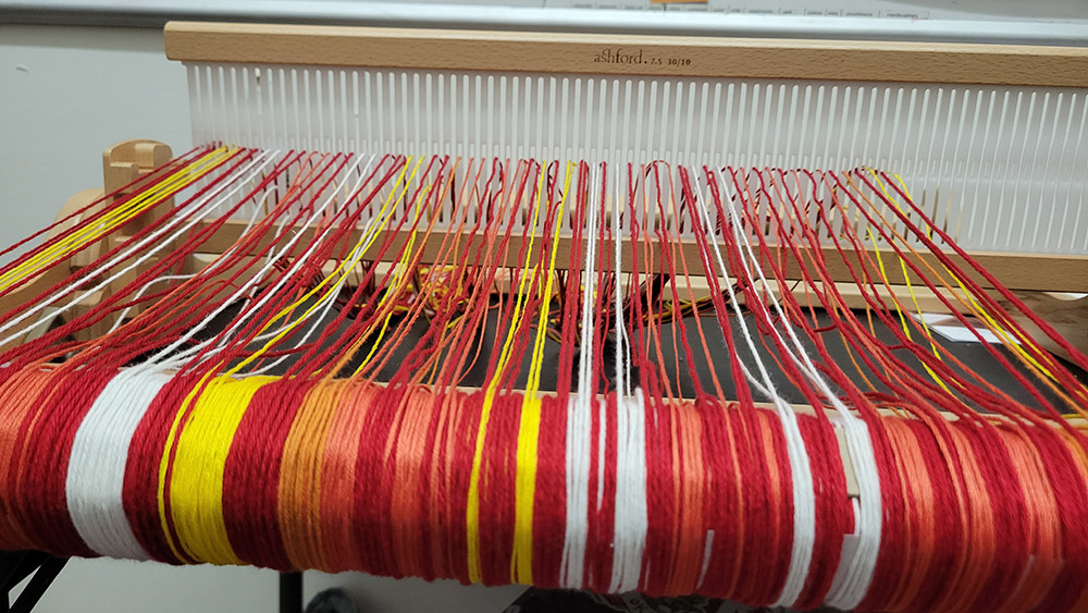 Warp threads on a rigid heddle loom, in shades of red, yellow, white, and orange